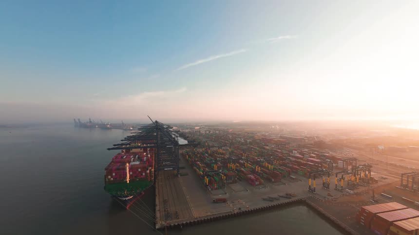 A sweeping aerial view reveals a massive container port with a cargo ship docked for loading as the sun rises in the background