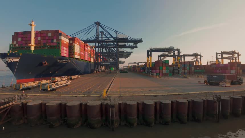 A massive container ship is docked at an industrial shipping port with cranes and containers during a hazy sunrise