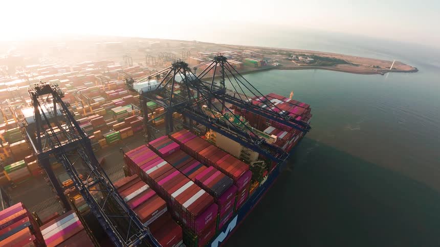 A cinematic aerial time-lapse shows massive gantry cranes loading a container ship at a bustling port during a hazy sunrise