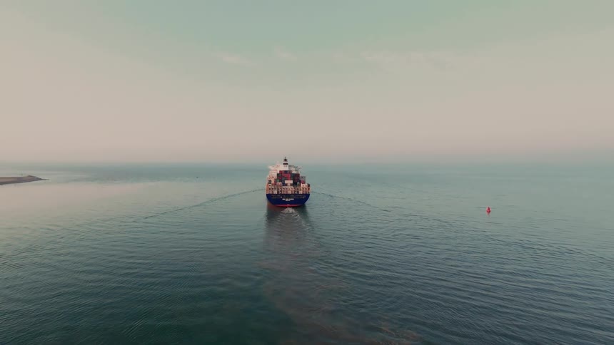 An aerial view follows a massive container ship as it sails through the calm, open ocean on a hazy day