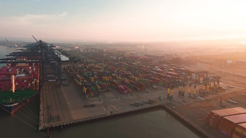 An expansive aerial shot reveals a massive shipping port at sunrise, with a large container ship docked and cranes lining the busy terminal