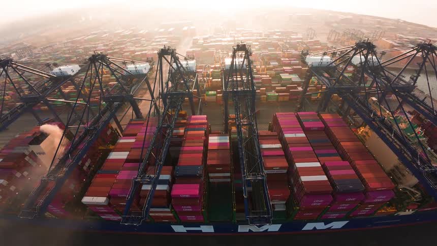 A wide-angle aerial time-lapse shows giant gantry cranes loading a massive cargo ship at a vast, hazy container terminal during a golden sunrise