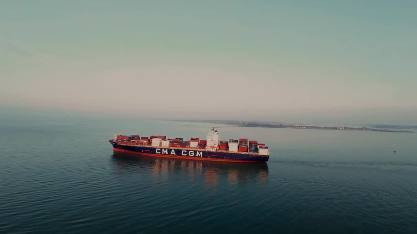 A cinematic aerial cinemagraph shows a large CMA CGM container ship sailing on the calm sea during a hazy day