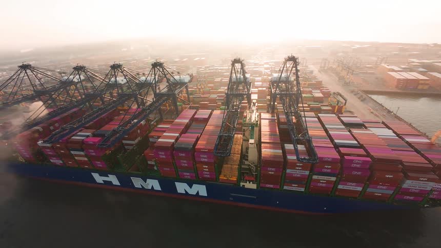 An aerial view shows a massive container ship being loaded by gantry cranes at a busy port during a hazy sunrise