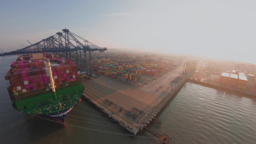 A wide aerial cinemagraph of a massive cargo container ship docked at a busy port during a hazy sunrise