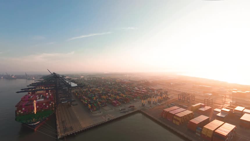 A stunning aerial view shows a massive container ship docked at a bustling commercial port filled with colorful shipping containers as the sun rises on the horizon