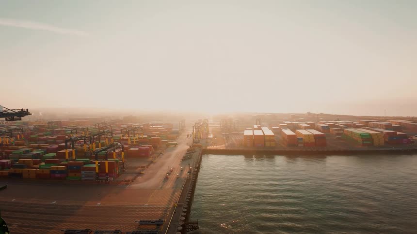 A vast shipping container port is busy with activity from cranes and vehicles during a hazy, golden sunset