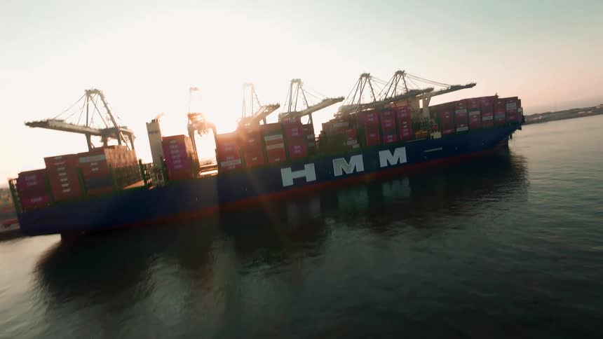 An aerial drone shot tilts up to reveal a massive container ship docked at a commercial port during a hazy sunrise