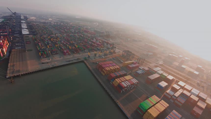 A sweeping wide-angle aerial shot captures a massive industrial shipping container terminal operating at sunrise