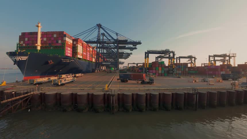 A static wide shot from the water shows a massive container ship docked at a bustling port while trucks move cargo during a beautiful sunrise