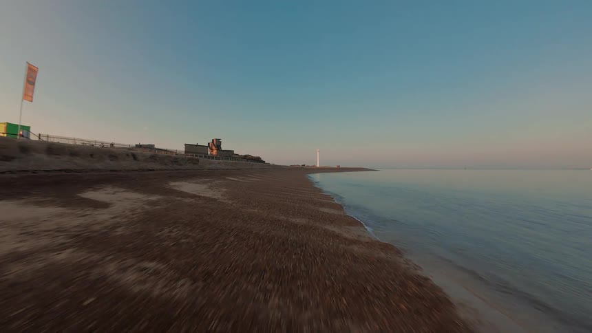A fast FPV drone flies low along a deserted shingle beach at sunrise, heading towards a distant lighthouse as a seagull takes off from the shore