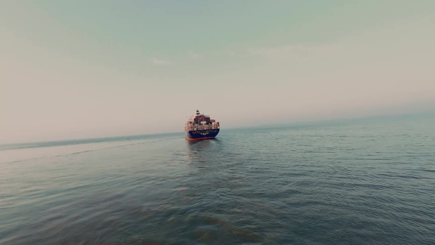 A large cargo container ship sails across the vast, calm ocean under a hazy sky