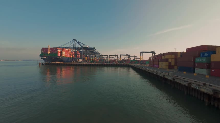 A massive container ship is docked at a bustling commercial port as cranes load and unload cargo during a beautiful sunrise
