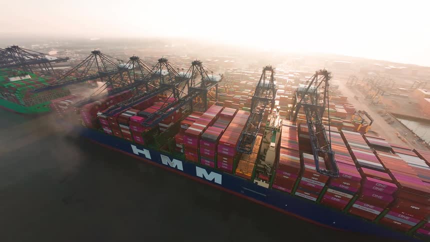An epic aerial view shows a massive container ship being loaded by gantry cranes at a bustling port during a hazy sunrise