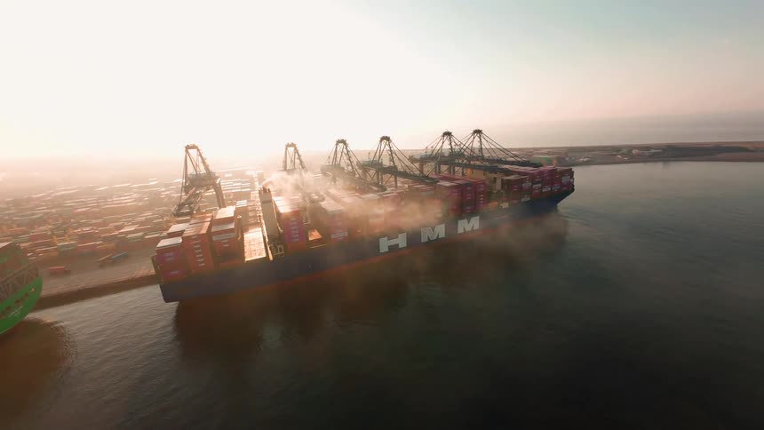 An epic aerial shot orbits a massive container ship being loaded by gantry cranes at a busy port during a foggy, golden sunrise