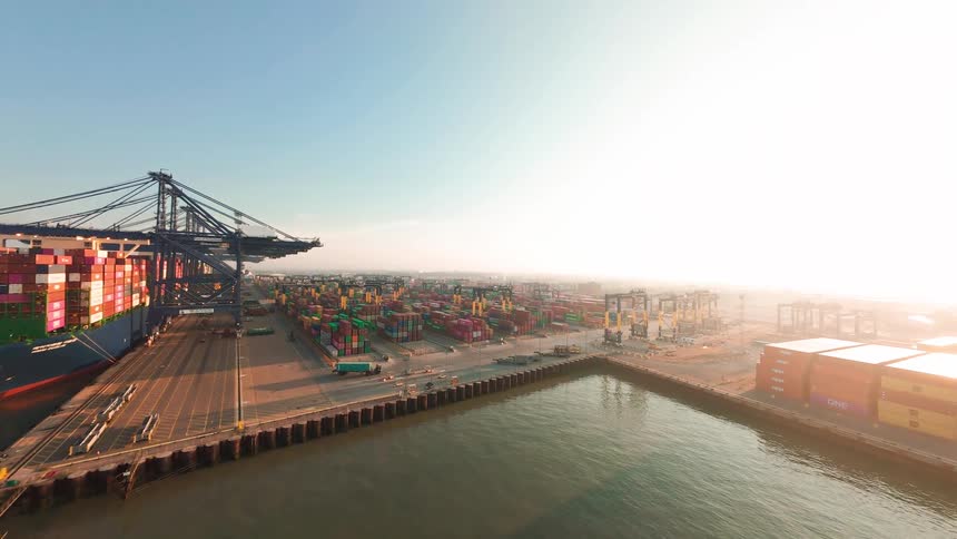 Expansive aerial shot of a large container ship docked at a bustling shipping port with cranes and thousands of containers during a bright sunrise