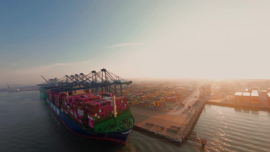 An aerial view shows a massive container ship being loaded by gantry cranes at a bustling port terminal during a golden sunrise