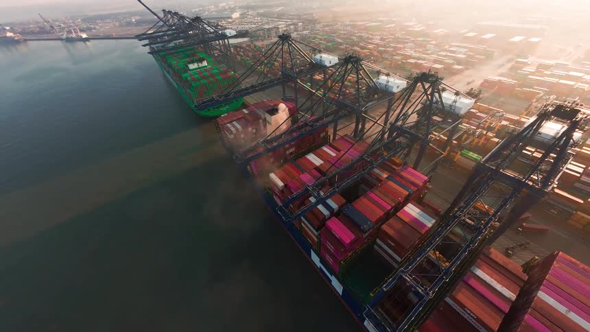 Aerial time-lapse of massive cranes loading and unloading colorful shipping containers from cargo ships at a bustling industrial seaport during a hazy sunrise