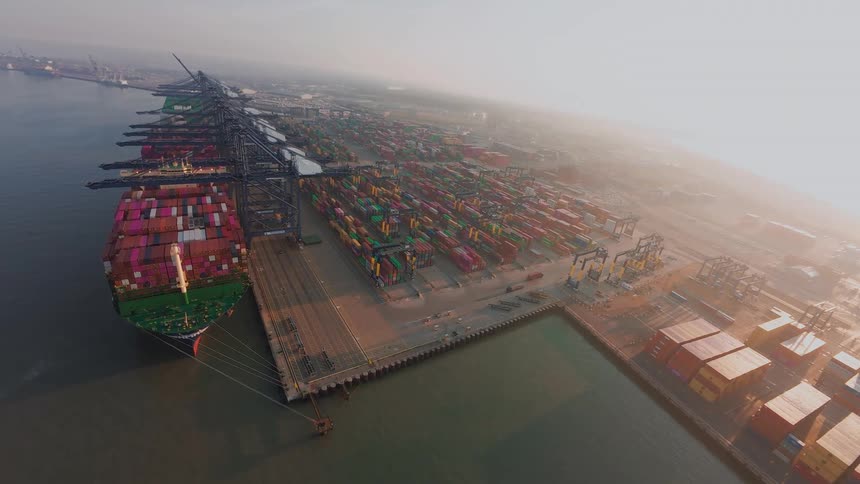 Aerial time-lapse of a large cargo ship being loaded and unloaded with shipping containers by gantry cranes at a busy port on a hazy day