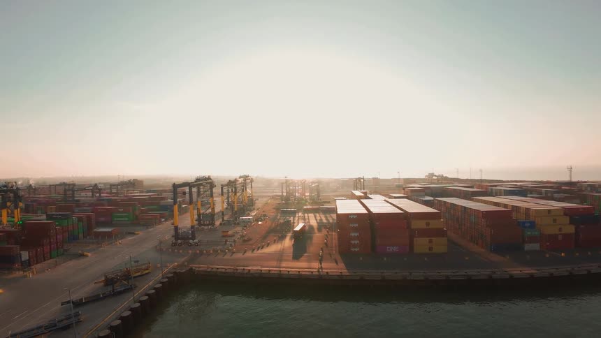A vast shipping container port comes to life with moving cranes and trucks under the warm glow of a hazy sunrise