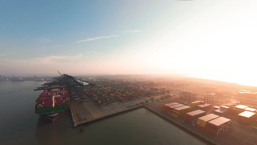 An expansive aerial view of a bustling industrial shipping port, where cranes load containers onto a massive cargo ship under a golden sunrise
