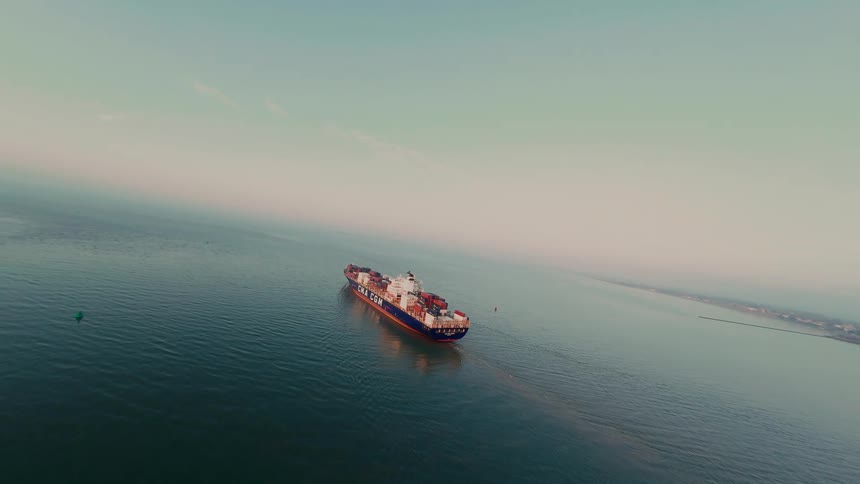 An expansive aerial view shows a large cargo container ship sailing across the calm open ocean under a hazy sky