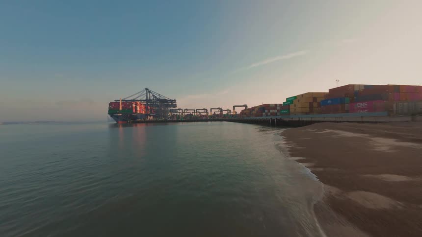 A low aerial flight over a sandy beach towards a large container ship docked at a commercial shipping port at sunrise