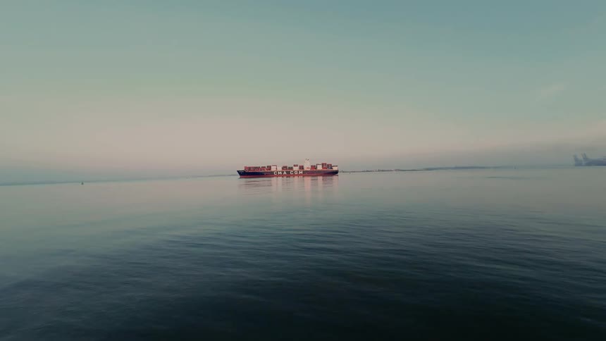 A wide, static shot of a large cargo container ship sailing across the calm ocean on a hazy morning