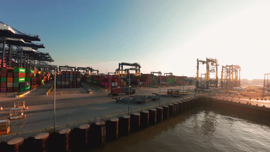 A slow aerial pan across a bustling industrial shipping port as the sun rises over the container yard and a docked cargo ship