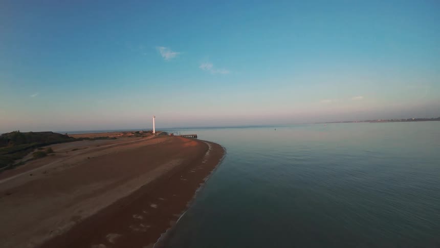 A serene aerial shot slowly pulling back from a lighthouse on a tranquil coastline during a beautiful golden hour