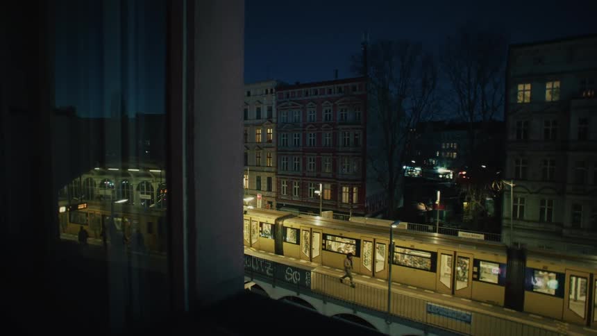 A yellow city train stops at an elevated station at night as passengers disembark before it departs, seen from a window view
