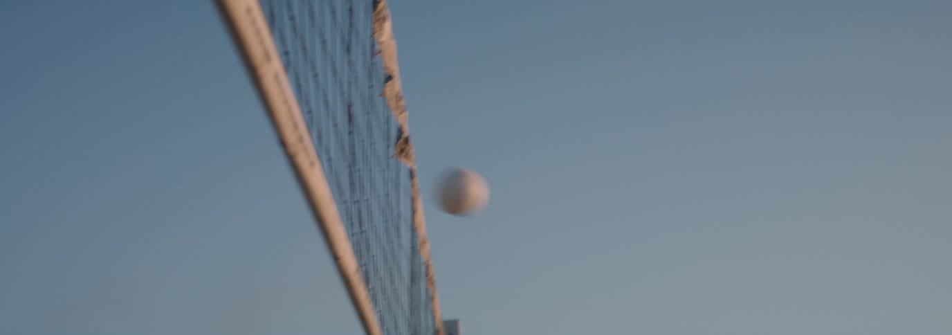 A man and woman in sunglasses play volleyball on the beach at dusk, ending the play with a high five