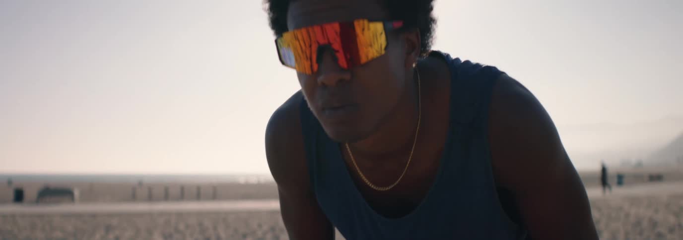 A determined young man in athletic sunglasses prepares to serve a volleyball on a sandy beach, tossing it up into the bright sun in slow motion
