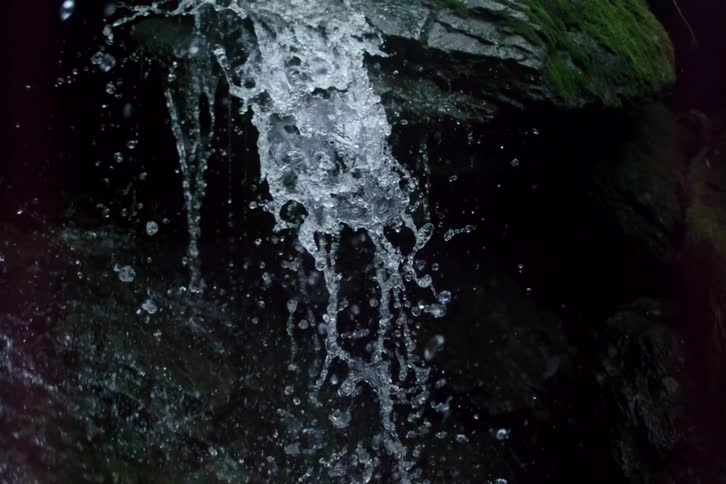 A close-up slow-motion shot of water from a stream cascading down dark, moss-covered rocks