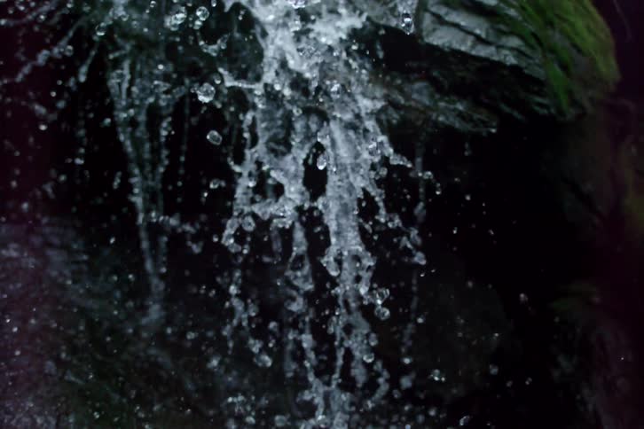A mesmerizing slow-motion close-up of water splashing against dark, moss-covered rocks in a waterfall