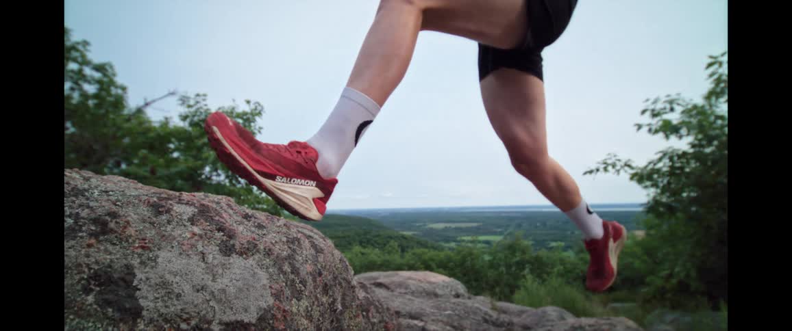 A low-angle cinemagraph captures the powerful leg of a trail runner mid-stride on a rock overlooking a valley