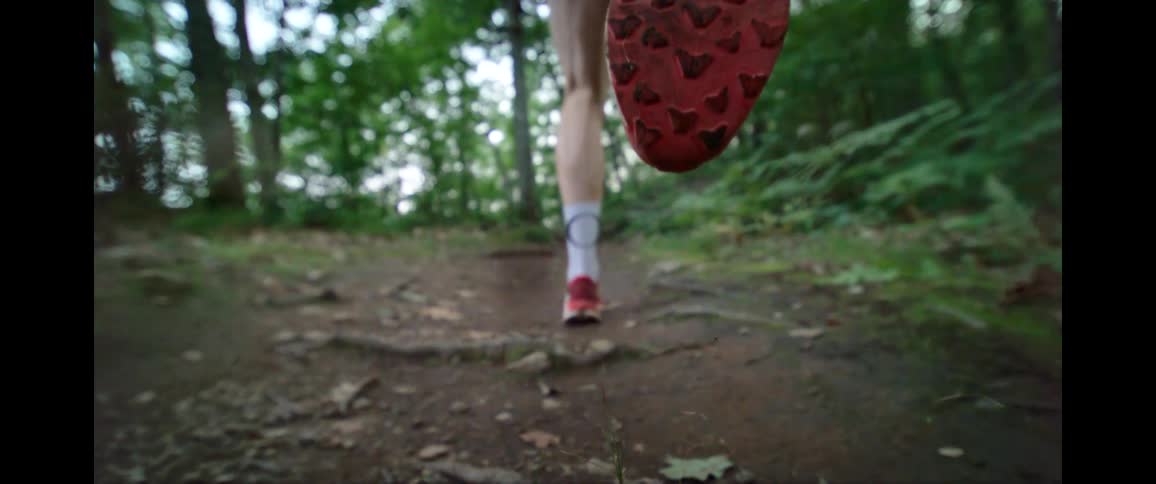 A super slow-motion close-up from behind of a runner's feet in red shoes as they jog along a dirt trail in the woods