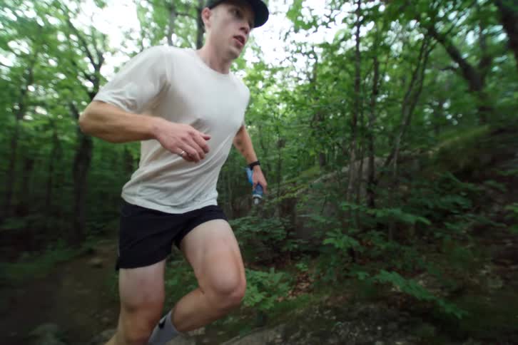 A determined male athlete trail runs up a steep, rocky path through a lush green forest in dramatic slow motion