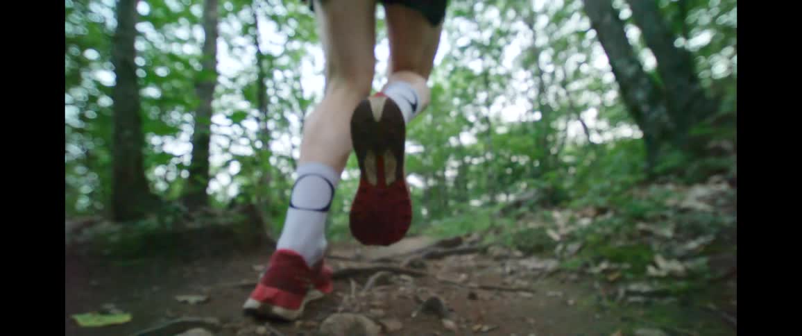 A super slow-motion, low-angle view of a runner's feet in red athletic shoes as they jog along a dirt trail in the woods