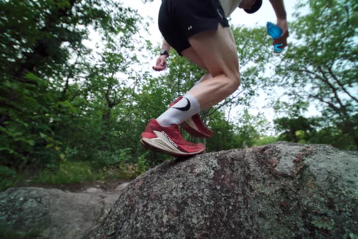 Super slow motion low-angle shot of a trail runner's muscular legs stepping onto a large rock in a forest