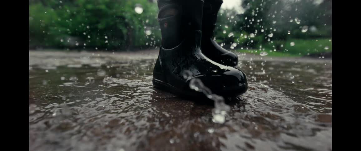Close-up of a person's black rain boots stepping into a puddle and creating a dramatic splash in super slow motion