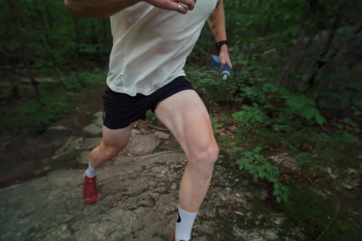 A determined male athlete holds a water bottle while running on a rocky trail through a lush green forest in slow motion