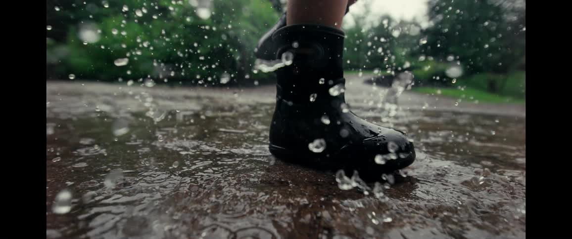 A dramatic super slow-motion close-up captures a person in rain boots stomping and splashing in a puddle on a rainy day