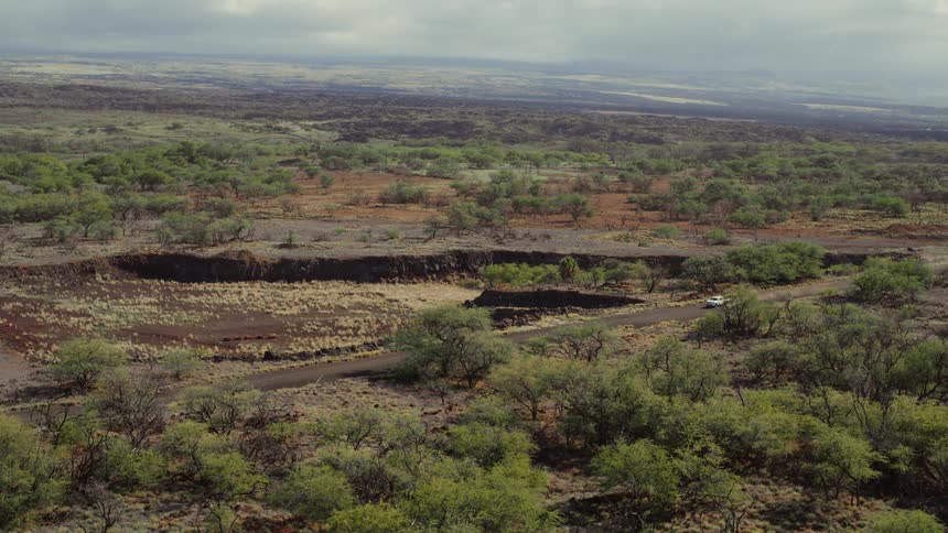 A white car drives on a remote dirt road through a vast volcanic landscape on an overcast day