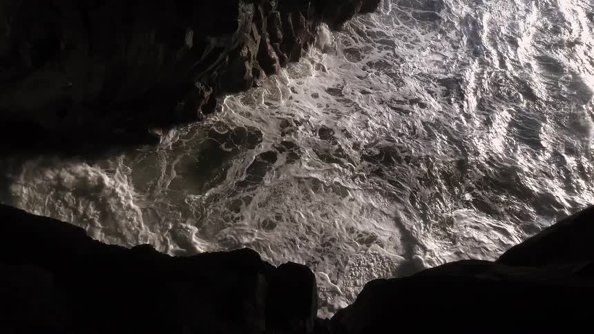 A powerful ocean wave crashes into a dark, rocky sea cave in dramatic slow motion