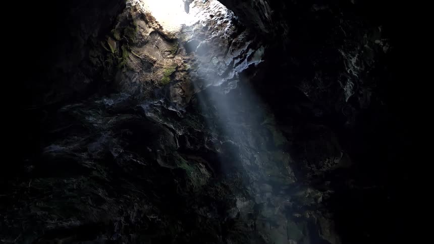 A beautiful sunbeam pierces through a hole in the ceiling of a dark, rocky cave, illuminating floating dust particles