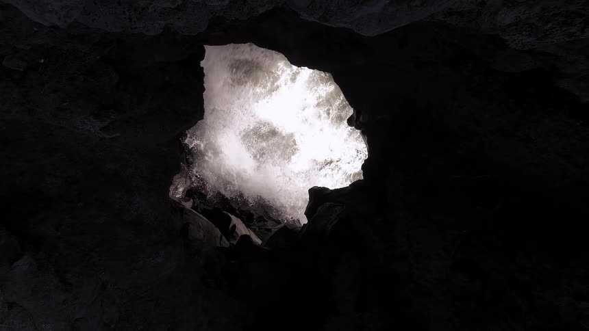 A dramatic slow zoom-out from inside a dark cave reveals a powerful waterfall crashing down from an opening above