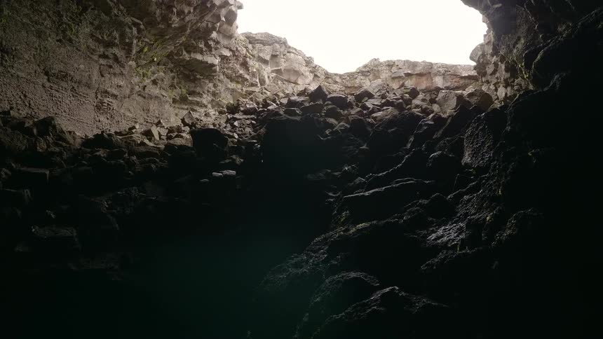Tilting up from the dark, rocky interior of a volcanic cave to reveal the bright sky through the entrance