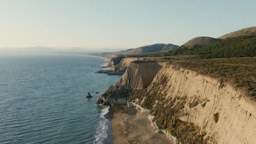 A static aerial view captures the stunning coastal landscape with towering cliffs meeting the ocean at sunset