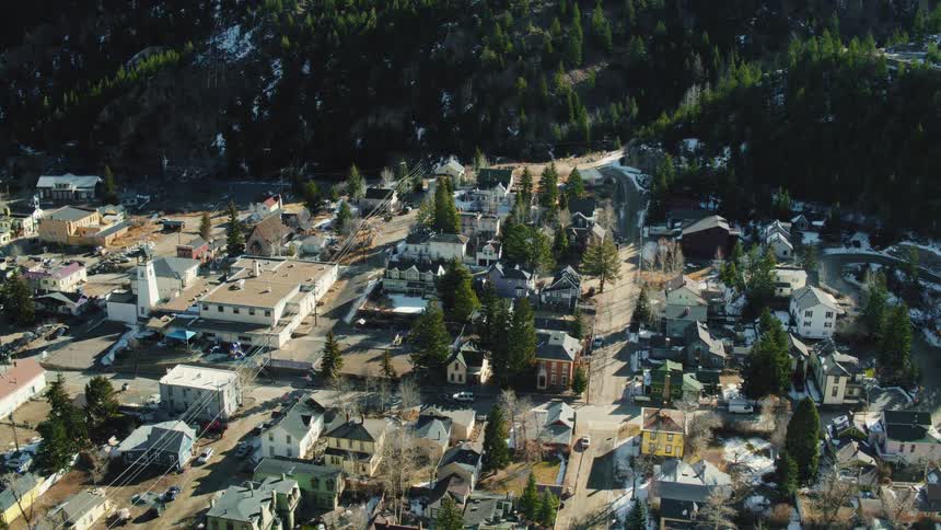 Aerial footage slowly zooms out over a quaint small town surrounded by forested mountains with patches of snow